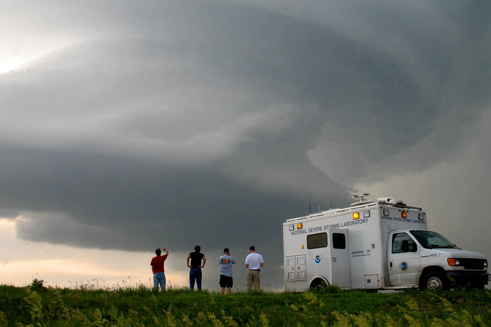 NSSL Command Vehicle in front of a thunderstorm cloud. Four researchers stand beside it, looking at the cloud.