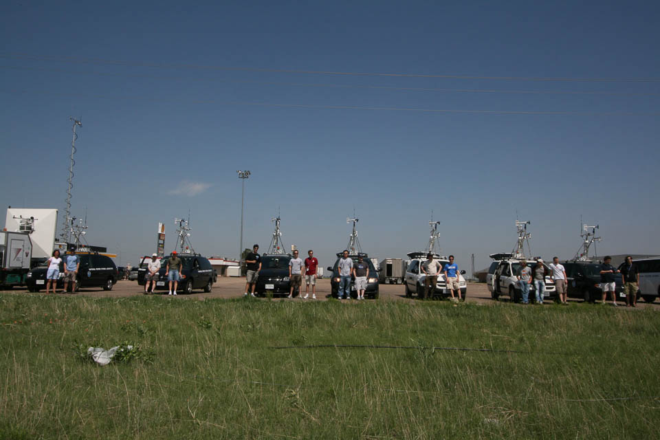 Multiple vehicles parked in a row in front of the camera. They have weather observing equipment mounted to the roofs. People are standing in front of each vehicle.