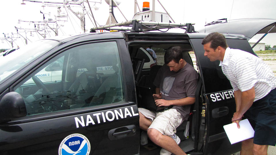 Two researchers confer over a laptop. One is sitting inside a minivan with a NOAA logo and 'National Severe Storms Lab' on the side; the other stands next to him